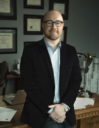 Andrew Carlton: Photograph of a man wearing a black blazer, light blue shirt, and wristwatch standing in an office setting. Background includes framed certificates on a wall, a wooden desk with papers, and architectural models or blueprints.